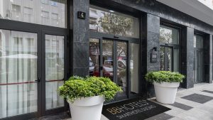 Entrance of BB Hotels Smart Hotel Duomo with glass doors, a black marble facade, large potted plants on either side of the entrance, and a logo mat on the sidewalk.