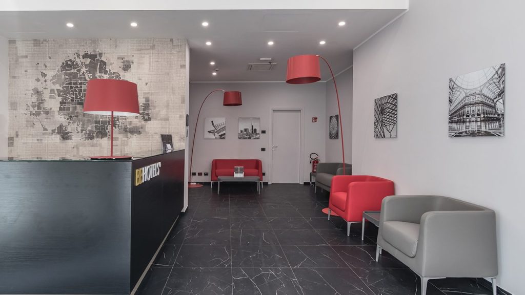 Modern office lobby with black reception desk, red and gray chairs, red floor lamps, black tile floor, abstract wall art, and minimalist decor. White walls with black-and-white framed photos complete the clean, contemporary space.