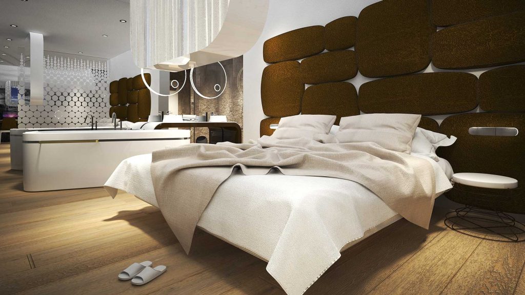 Modern hotel room with a large bed covered in white linens, two pillows, and a light blanket. Brown padded headboard, wooden floor, slippers, and a white freestanding bathtub in the background. Contemporary decor.