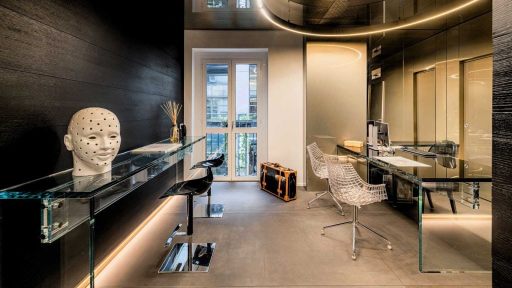 A modern office with glass desks, transparent chairs, black bar stools, a decorative head sculpture, and a large window letting in natural light. A suitcase is on the floor near a reflective wall.