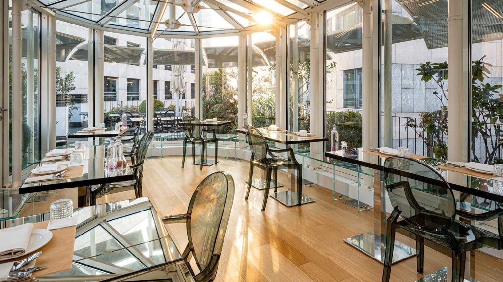 A bright, modern restaurant dining area with glass walls and ceiling, clear chairs, glass tables, and sunlight streaming in. Place settings and glassware are arranged neatly on the tables.