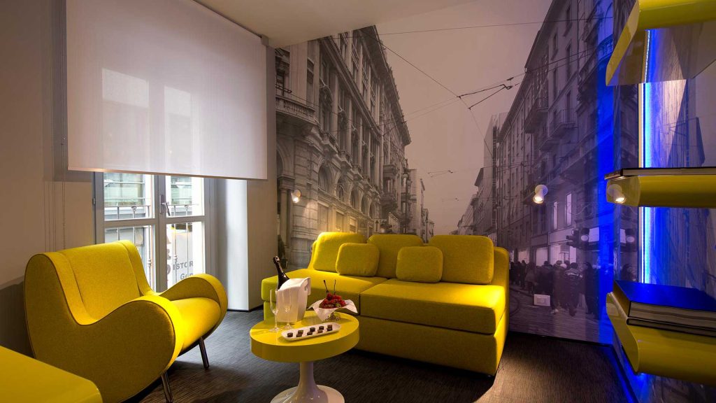Modern living room with yellow furniture, a round table with snacks and champagne, a large wall mural of a city street, and shelves with blue accent lighting. Natural light streams in through a window with a white blind.