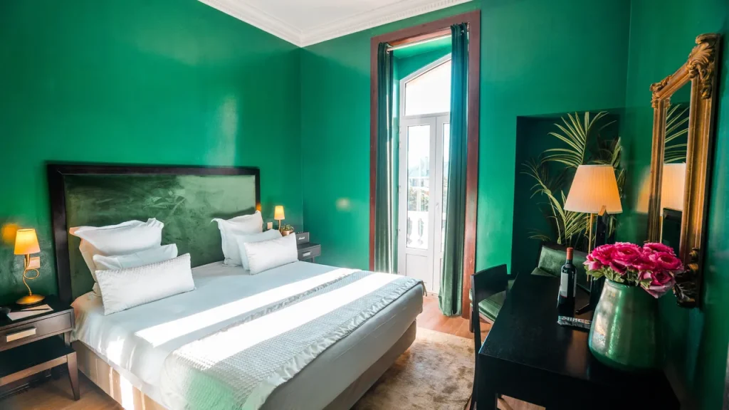 A bright, stylish bedroom with green walls, a large bed with white linens, a dark wood desk with a lamp and pink flowers, and tall windows letting in natural light.