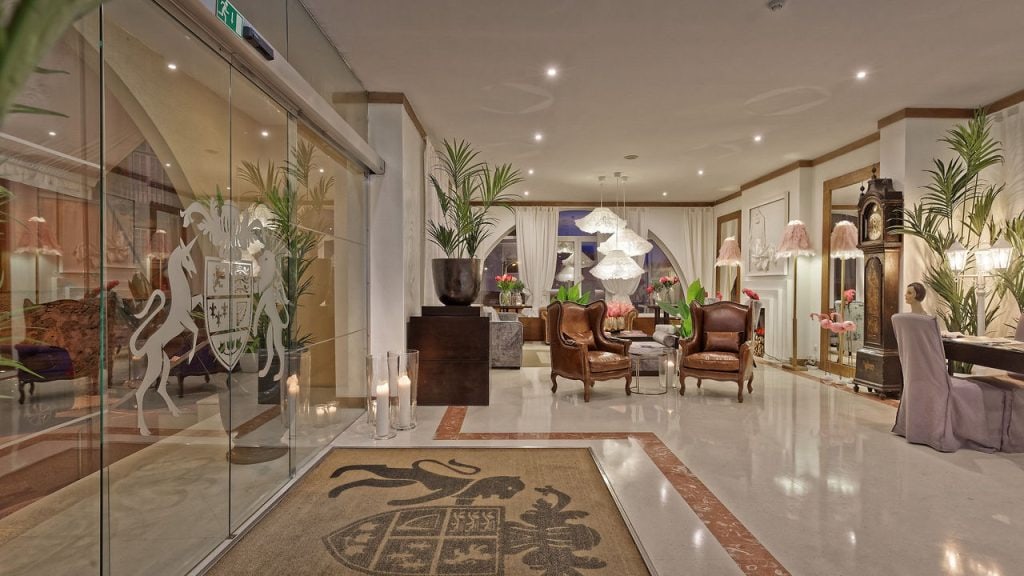 Elegant hotel lobby with glass doors, ornate rug, armchairs, tall plants, modern chandeliers, a grandfather clock, and soft lighting. The decor mixes classic and contemporary furnishings in a spacious, inviting setting.