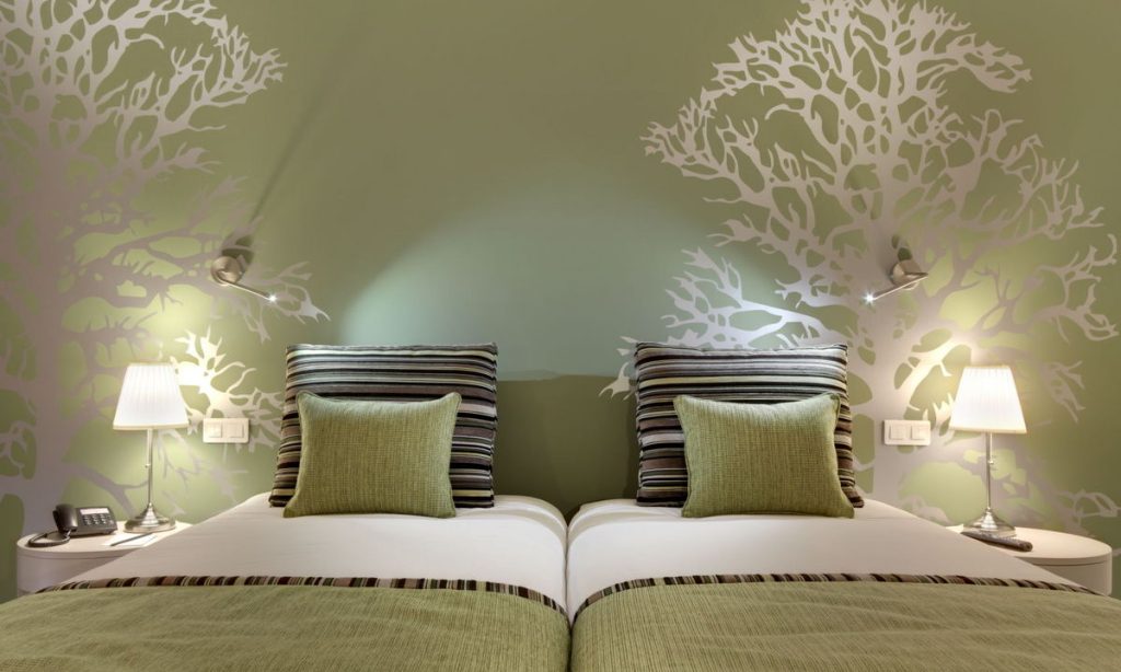 Modern bedroom with two twin beds, striped pillows, green bedspreads, wall-mounted reading lights, bedside tables with lamps, and a green wall featuring white tree silhouette decals.