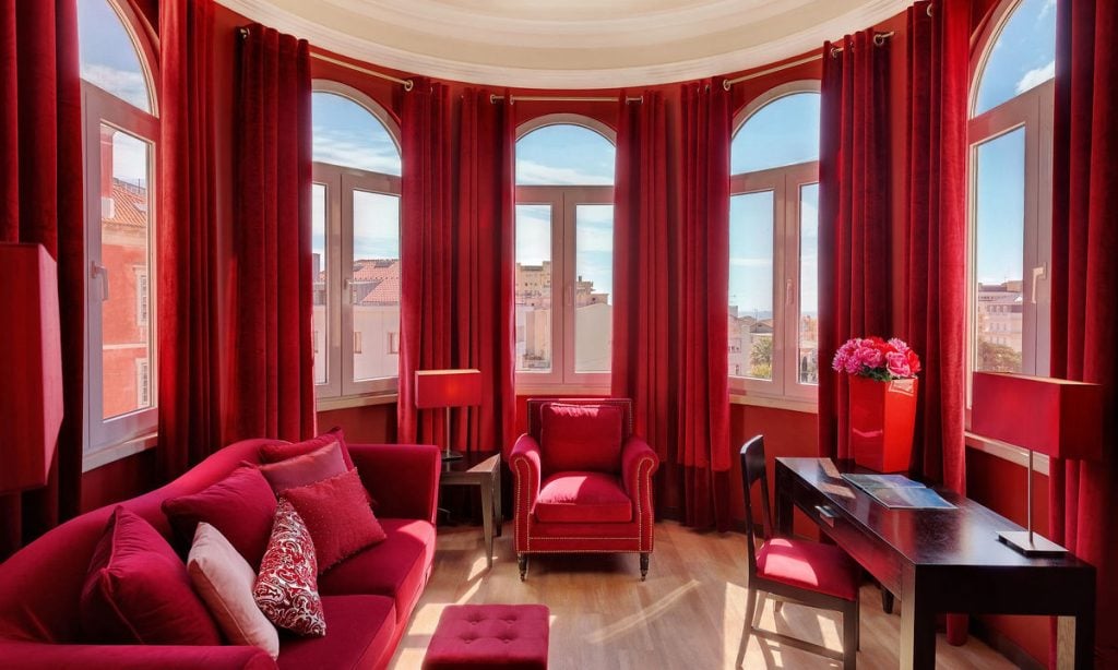 A bright room with tall arched windows and red curtains, featuring a red sofa, armchair, ottoman, desk, and lamps. Pink flowers in a vase and decorative pillows add to the vibrant decor.