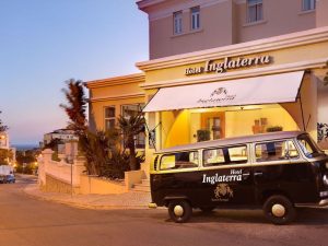 A black van with Hotel Inglaterra parked outside a beige hotel with the same name, featuring an awning and warm lights at sunset on a quiet street.