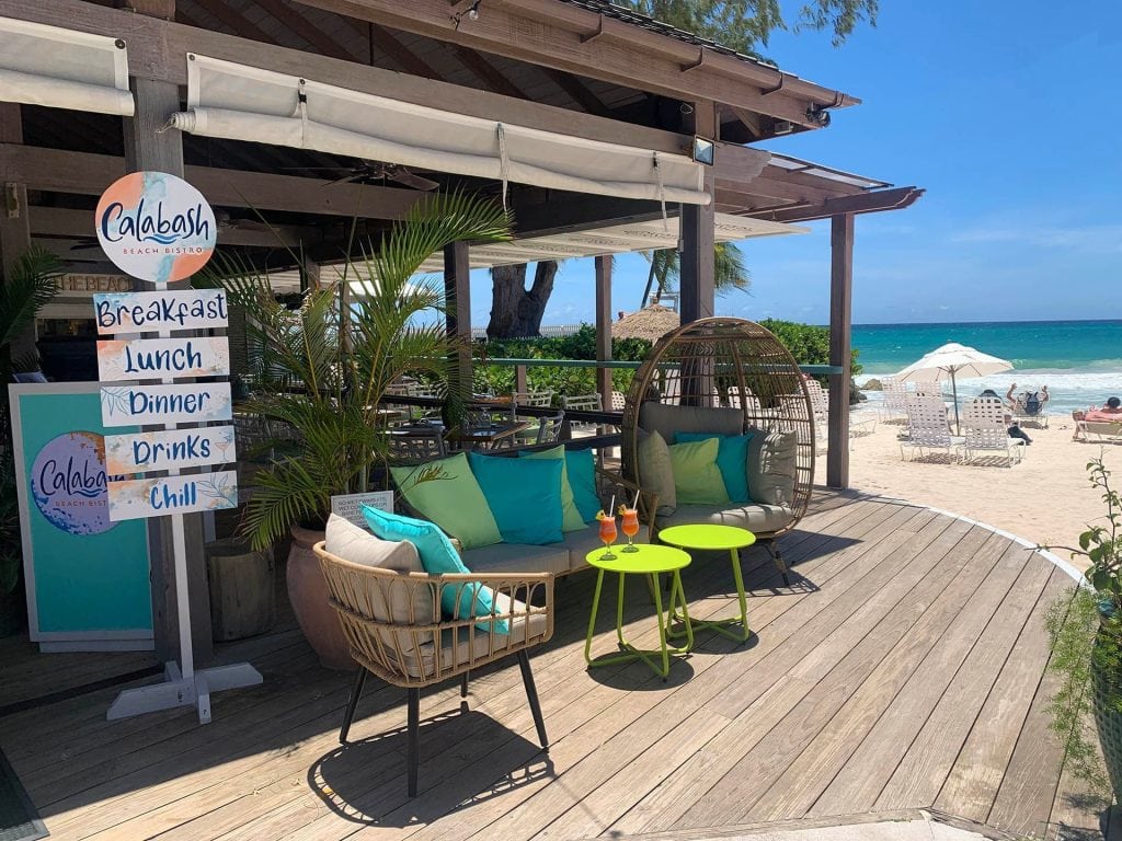 Outdoor beachside lounge area with cushioned chairs, a small table holding two drinks, and a sign listing Breakfast, Brunch, Dinner, Drinks, Chill. The sandy beach and ocean are visible in the background.
