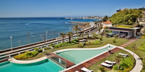 A curved outdoor pool with lounge chairs and gardens overlooks a calm blue sea, marina, and coastal town skyline. Palm trees and a modern poolside building enhance the relaxing resort atmosphere.