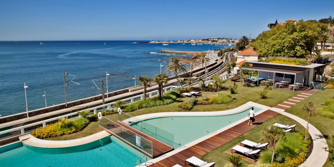 A curved outdoor pool with lounge chairs and gardens overlooks a calm blue sea, marina, and coastal town skyline. Palm trees and a modern poolside building enhance the relaxing resort atmosphere.