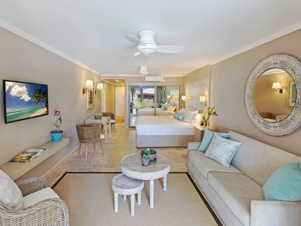 A bright, airy hotel room features a large bed, sofa, wicker chairs, round coffee tables, a wall-mounted TV, and a large decorative mirror. Soft neutral colors and tropical accents create a relaxing, beach-inspired atmosphere.