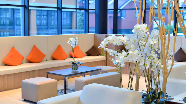 Modern lounge with a beige sectional sofa, orange accent pillows, small square ottomans, a coffee table with a potted orchid, and large floor-to-ceiling windows letting in natural light. White flowers decorate the space.