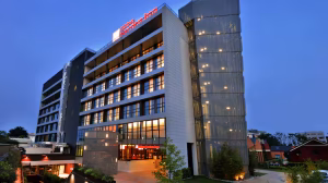 Modern multi-story hotel building, illuminated at dusk with warm lights in the windows, featuring a prominent sign reading “Hilton Garden Inn” at the top, surrounded by greenery and clear evening sky.