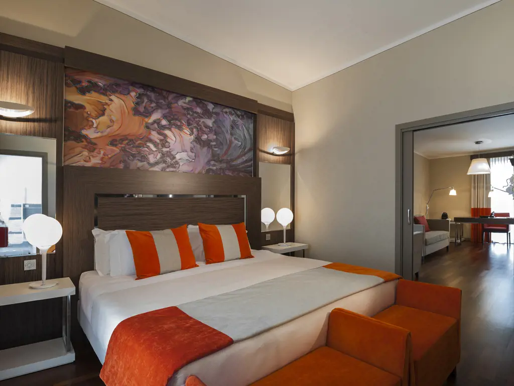 A modern hotel room with a large bed, orange and white bedding, abstract art above the headboard, two bedside tables with spherical lamps, a bench at the foot of the bed, and an adjacent sitting area visible through an open doorway.