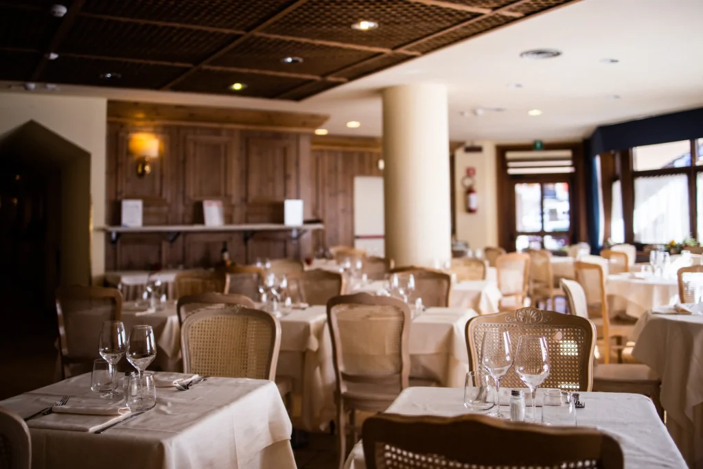 A cozy restaurant dining area with neatly set tables covered in white tablecloths, each arranged with glassware and cutlery. The room has wooden accents, soft lighting, and empty chairs, creating an inviting atmosphere.