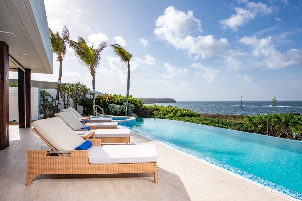 A modern infinity pool with wicker lounge chairs overlooks the ocean, bordered by lush greenery and palm trees, under a partly cloudy sky.