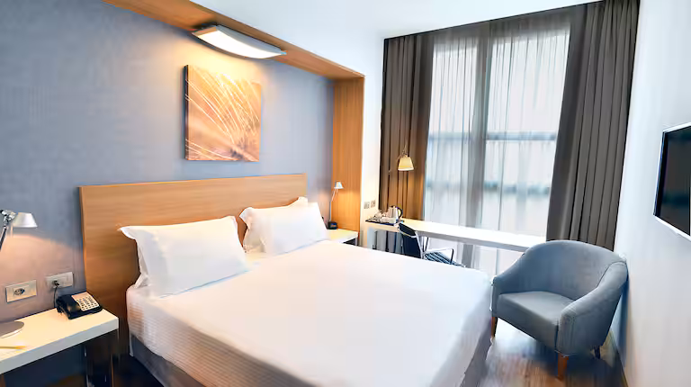 Modern hotel room with a double bed, light wood headboard, two white pillows, desk with a lamp, armchair, wall-mounted TV, and large window with sheer and dark curtains. Warm, minimalist decor.