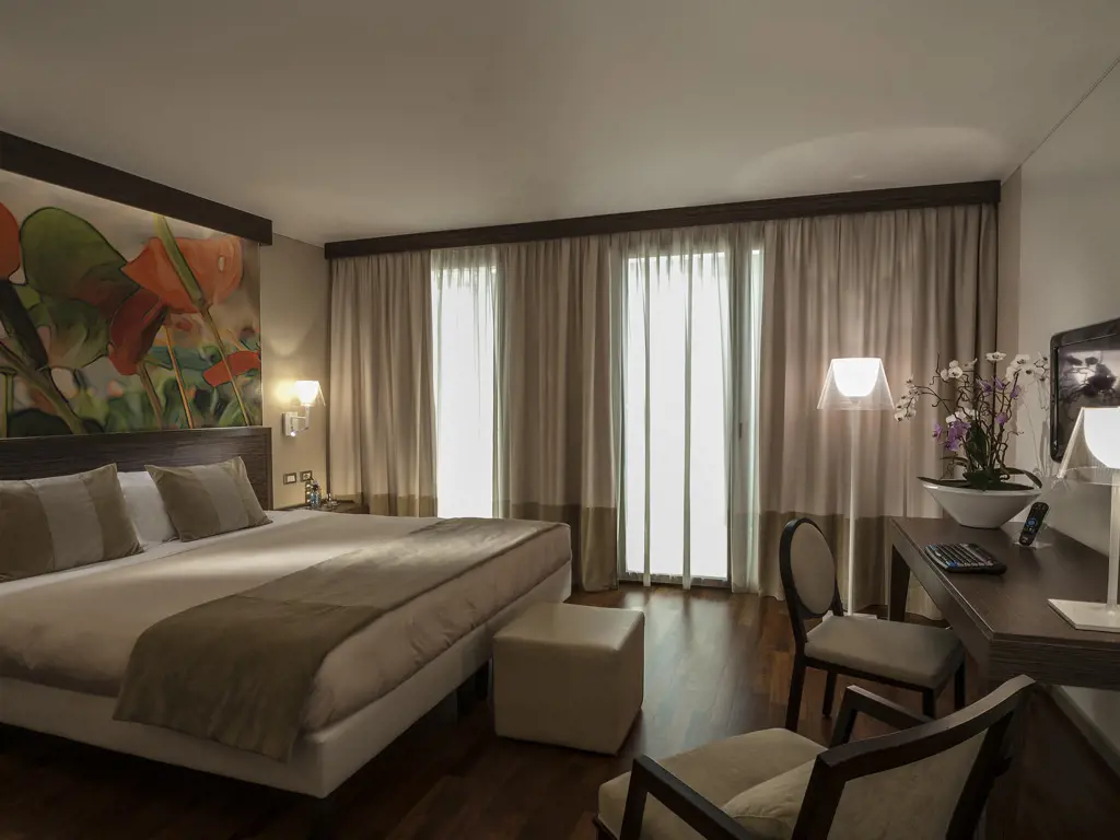 A modern hotel room with a large bed, neutral bedding, a floral wall mural, two armchairs, a desk with a chair, a TV, and floor-to-ceiling curtains covering windows letting in natural light.