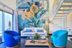Modern living room with a white sofa, two blue armchairs, a glass coffee table, pendant lights, tropical wall mural, and white stairs. Large windows let in natural light. The decor is colorful and vibrant.