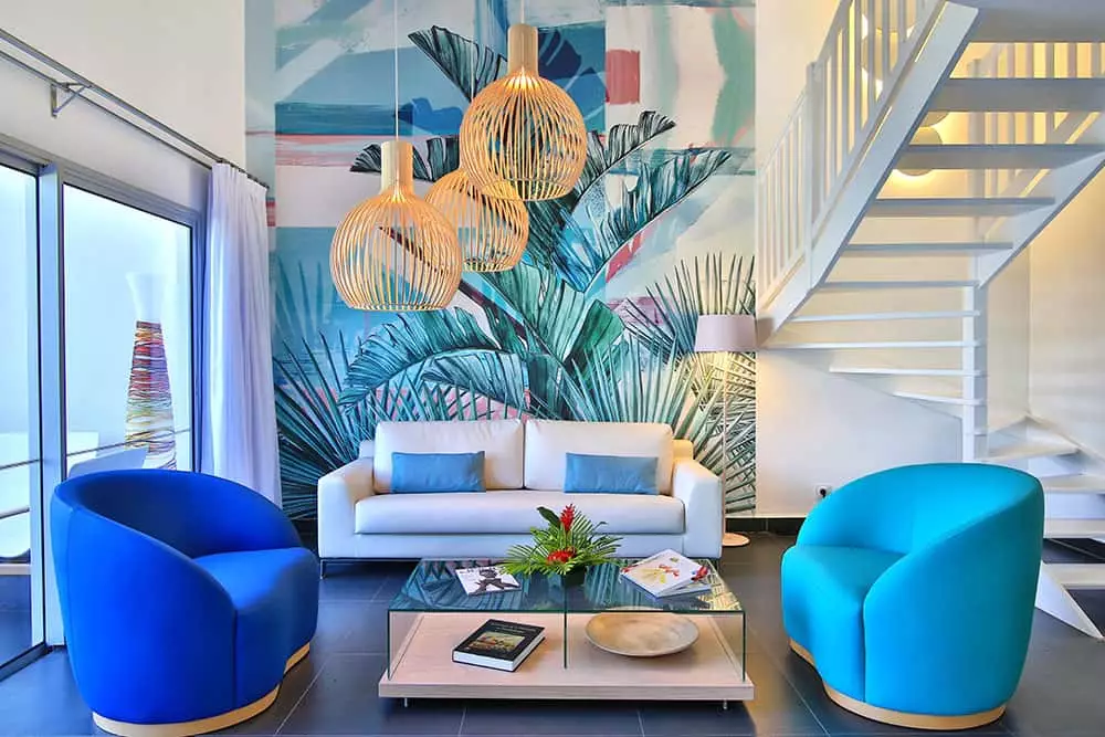 Modern living room with a white sofa, two blue armchairs, a glass coffee table, pendant lights, tropical wall mural, and white stairs. Large windows let in natural light. The decor is colorful and vibrant.