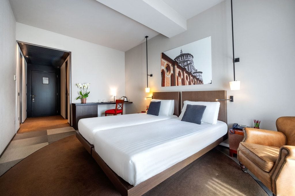 A modern hotel room with a large double bed, white linens, dark headboard, two pillows, a desk with a red chair, an armchair, and a photo of a historic building on the wall. Bright, minimalist decor.
