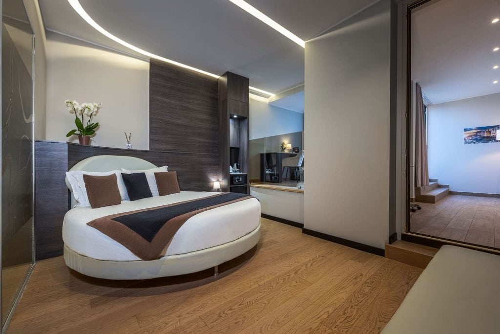 Modern hotel room with a round bed, brown and white bedding, wood paneling, orchid plant, TV, and glass partition. Room features minimalist decor and warm lighting, with a hallway visible through an open door.