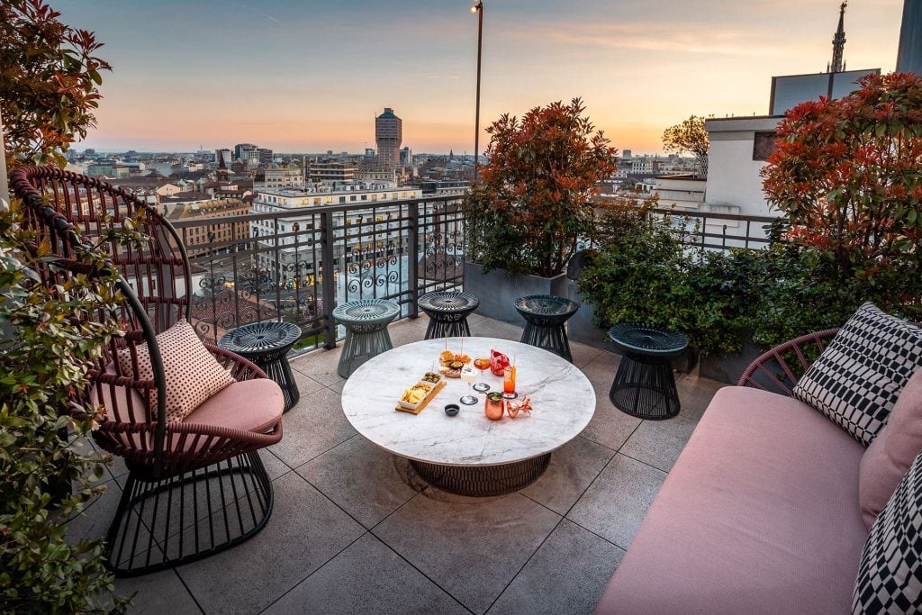A rooftop terrace at sunset with a round table set for snacks and drinks, surrounded by modern chairs and a sofa, overlooking a city skyline with buildings and a distant tower. Shrubs decorate the edges of the terrace.