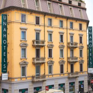 A yellow building with UNAHOTELS signs on both corners, featuring balconies, shuttered windows, and shops at street level. The entrance is visible, along with a red metro sign in front.