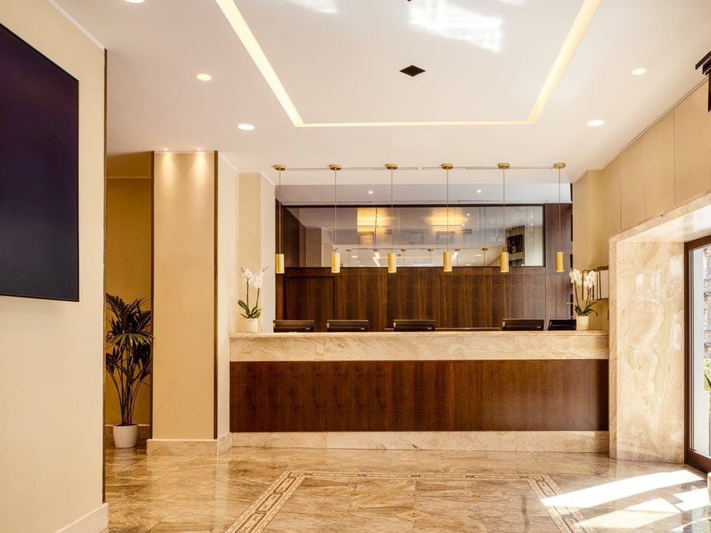A modern hotel reception area with a marble front desk, wood paneling, pendant lights, potted plants, and orchids. The floor is polished marble, and large windows let in natural light.