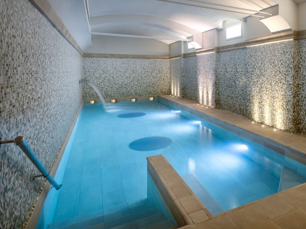 A modern indoor spa pool with mosaic tiled walls, underwater lighting, and a small fountain feature. The pool is rectangular with steps and a handrail, creating a relaxing and luxurious atmosphere.