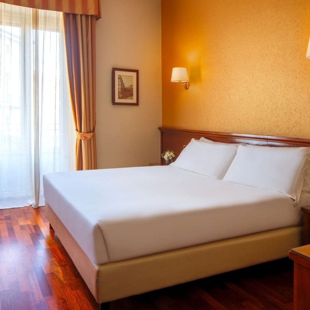 A bright hotel room with a double bed, white linens, wooden headboard, yellow wall, wall lamps, framed artwork, small bedside table with flowers, and sunlight streaming through sheer curtains.