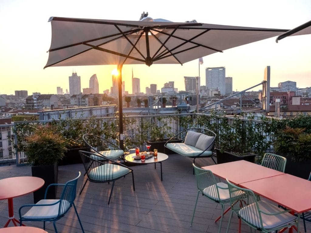 A rooftop terrace with modern tables and chairs, potted plants, and a large umbrella overlooks a city skyline at sunset. Drinks and snacks are set on a table, creating a relaxed, inviting atmosphere.