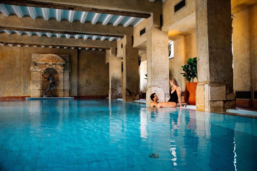Two women relax by the edge of an indoor swimming pool with classical architecture, stone columns, and a statue in the background. Warm lighting and large potted plants create a serene atmosphere.