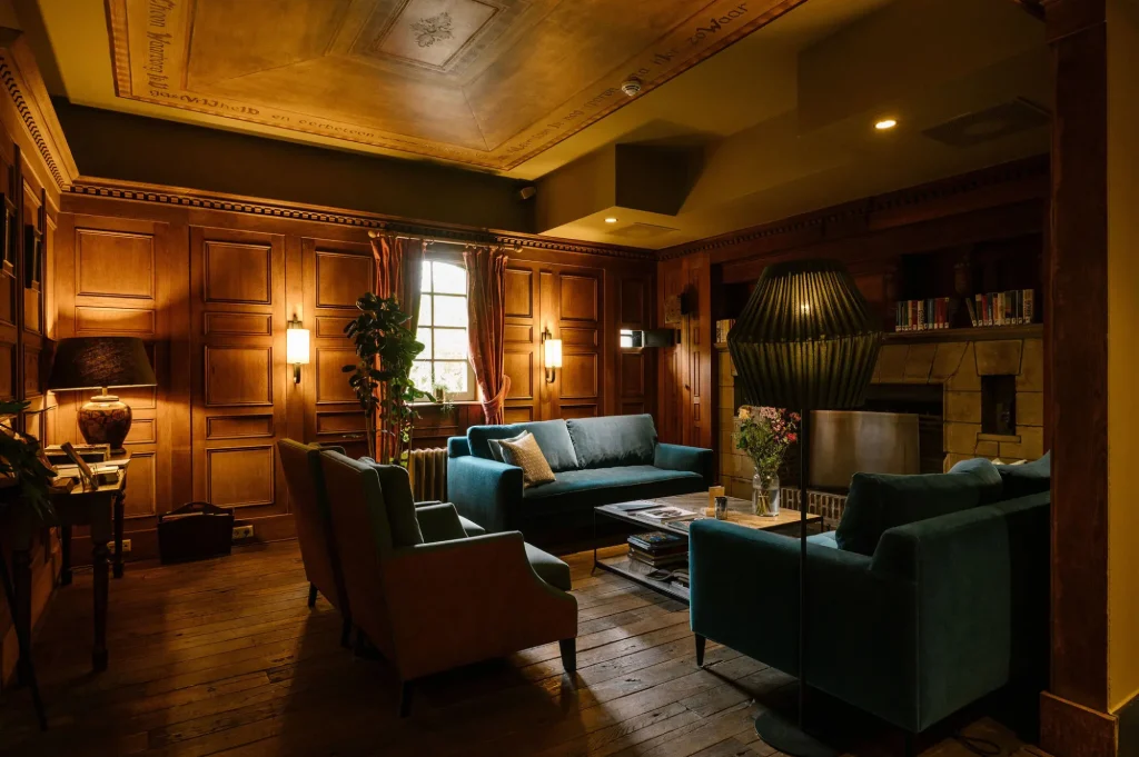 A cozy, warmly lit lounge features teal sofas, an armchair, wood-paneled walls, a fireplace, bookshelves, and a large black floor lamp, creating an inviting atmosphere with vintage decor and wooden flooring.