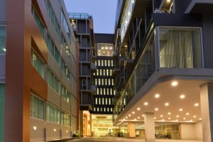 Modern multi-story office buildings with large windows and bright exterior lighting; the photo is taken at dusk, showing an open area between the structures and illuminated interior spaces.