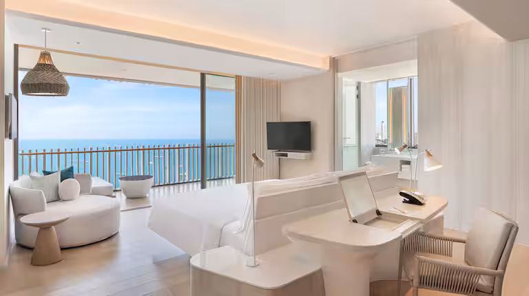 Modern hotel room with a large bed, desk, chair, flat-screen TV, seating area, and floor-to-ceiling windows opening to a balcony with ocean views under a clear blue sky. Decor is light and minimalist.