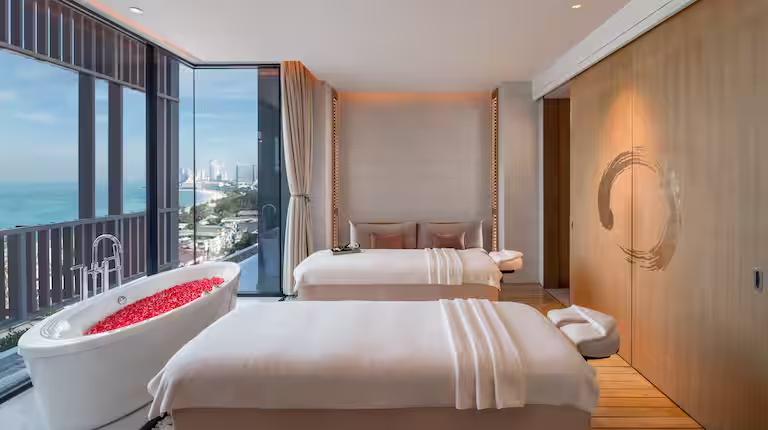 A modern spa room with two massage tables, a bathtub filled with red flower petals, floor-to-ceiling windows offering an ocean view, and minimalist decor with wood and beige tones.