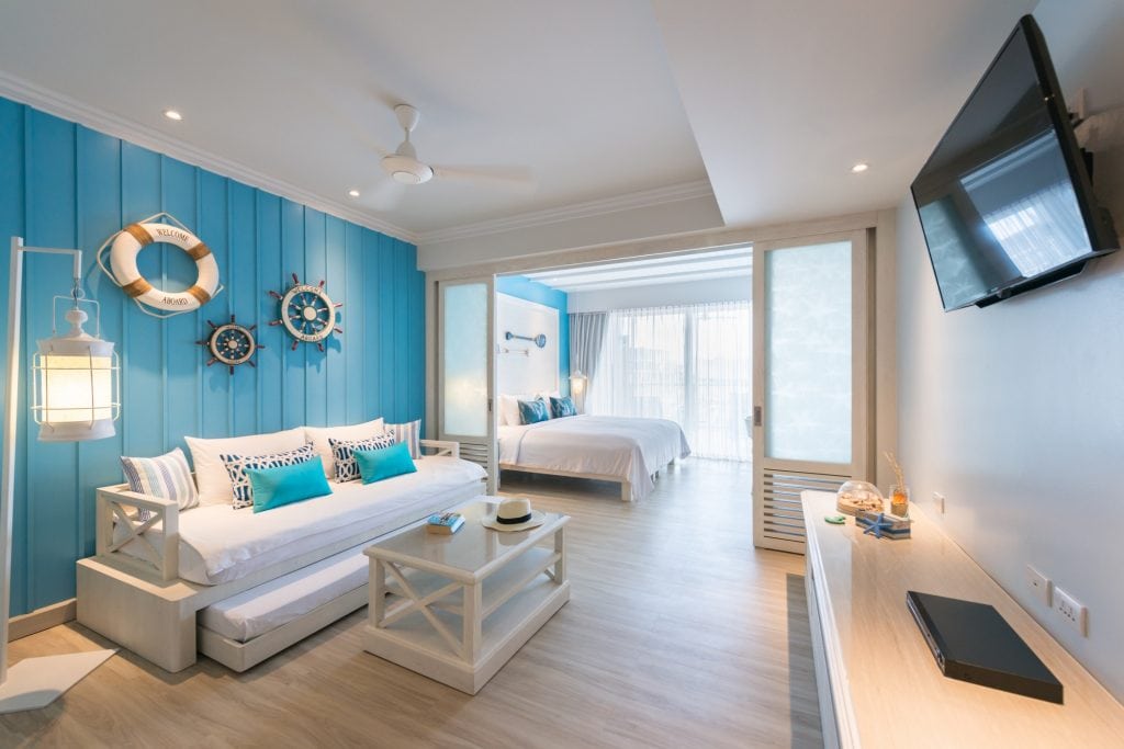 A bright, nautical-themed hotel room with blue accent wall, white furniture, a large bed, sofa, coffee table, TV, and nautical decor such as lifebuoys and ship wheels. Sunlight streams in through large windows.