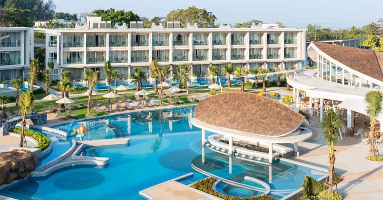 Aerial view of a modern resort with a large, curved swimming pool, poolside bar with a thatched roof, lounge chairs, umbrellas, palm trees, and a multi-story hotel building in the background.