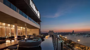 Outdoor seating area with modern round sofas and tables beside a reflective pool on a high-rise Hilton hotel balcony, overlooking a city skyline and river at sunset.