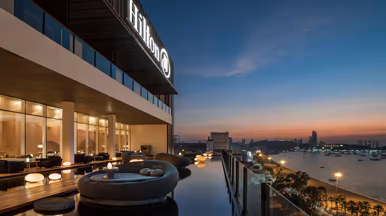Outdoor seating area with modern round sofas and tables beside a reflective pool on a high-rise Hilton hotel balcony, overlooking a city skyline and river at sunset.