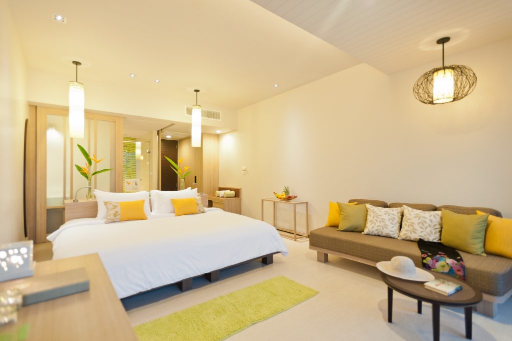 A bright, modern hotel room with a large bed, neutral walls, colorful yellow and green pillows, a sofa, two pendant lights, and tropical plants for decor. A straw hat is placed on the sofa.
