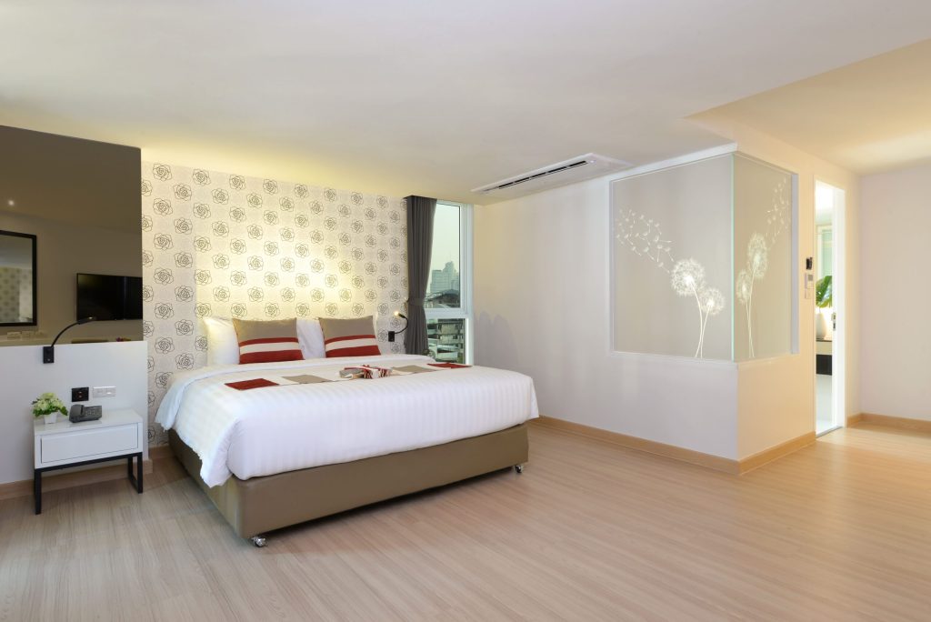 A modern hotel room with a large bed, white bedding, and decorative pillows. The room has light wood flooring, patterned wallpaper, a TV, a small bedside table, and a glass partition with dandelion designs.