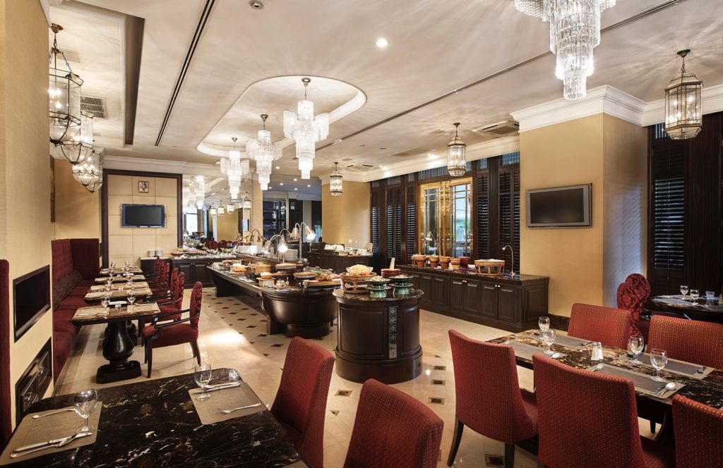 Elegant restaurant dining area with red upholstered chairs, set tables, chandeliers, and buffet stations filled with various dishes. The space features warm lighting, large mirrors, and luxurious decor.