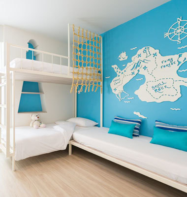 Children’s bedroom with a blue accent wall featuring a white pirate map, a white bunk bed with a climbing net, a teddy bear on the lower bunk, and a single bed with blue cushions.