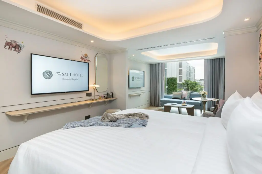 A modern hotel room with a large bed, wall-mounted TV, small sitting area by a window, and soft lighting. The room features white bedding, light walls, and a window with a city view and greenery outside.
