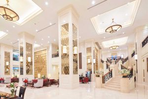 A luxurious hotel lobby with marble floors, tall decorative columns, elegant chandeliers, plush seating areas, floral arrangements, and a grand staircase with ornate railings leading to an upper level.