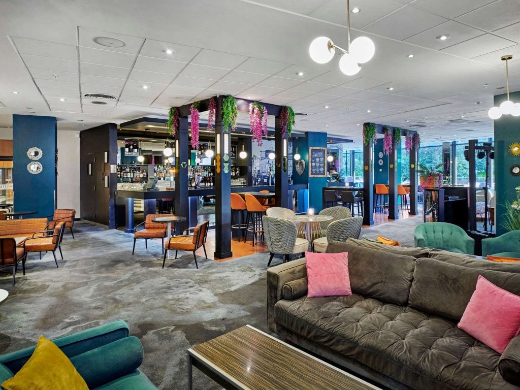 Modern hotel lounge with stylish furniture, including gray and teal sofas, pink and gold cushions, and chairs around small tables. The space features a bar, hanging plants, and decorative lighting.