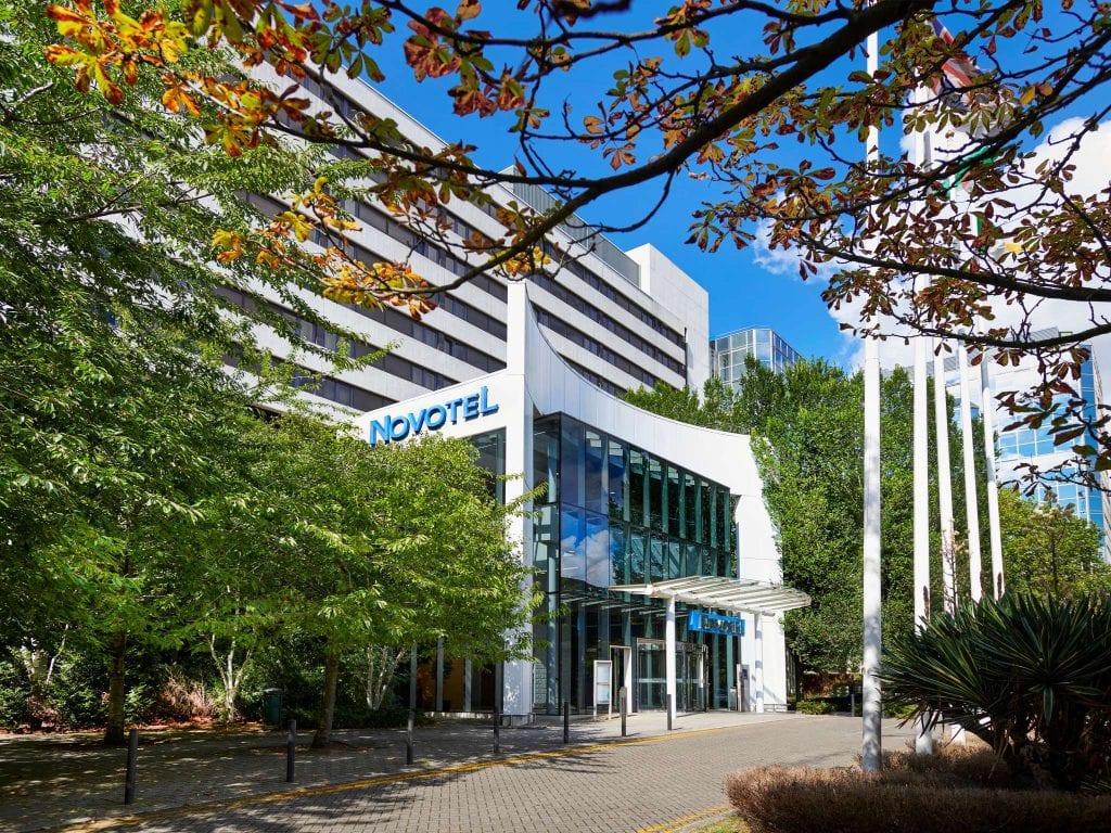 The exterior of a modern Novotel hotel building with large glass windows, surrounded by green trees and white flagpoles, under a blue sky with scattered clouds.