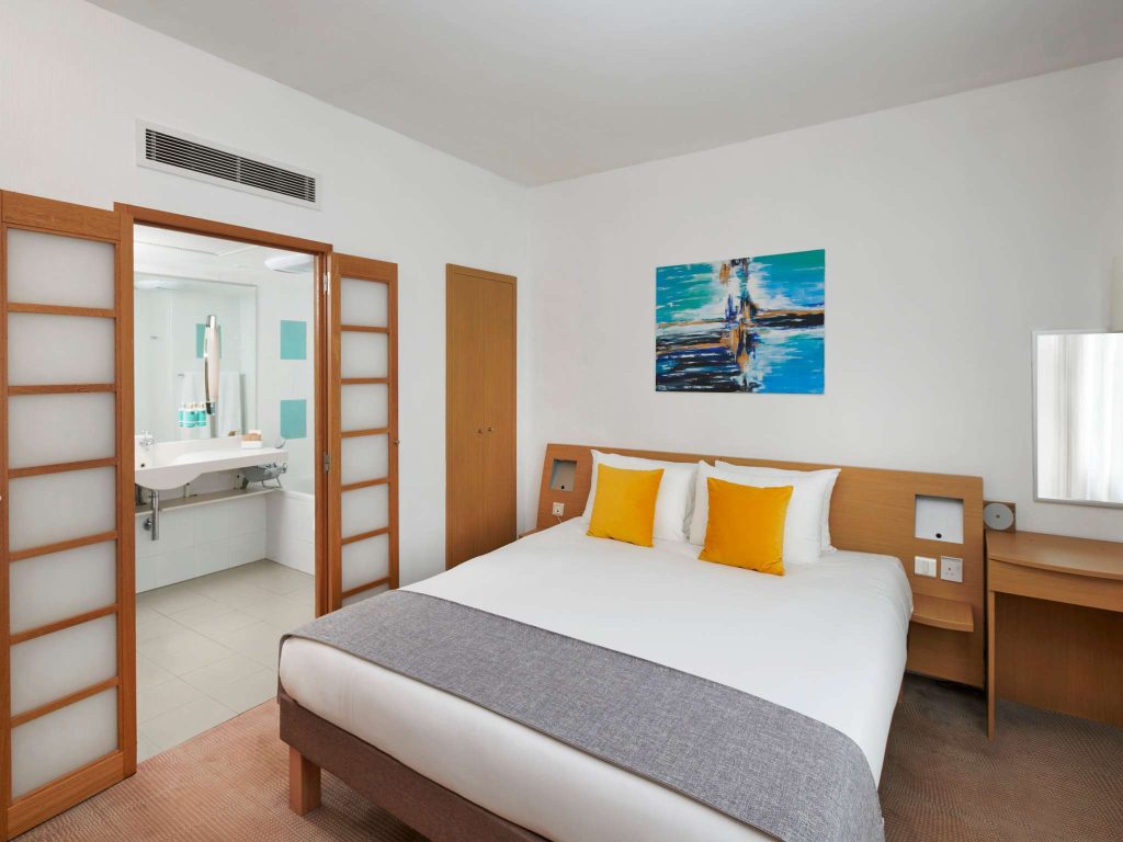 Modern hotel room with a large bed, two yellow accent pillows, a desk, and an open sliding door leading to a bright bathroom. Abstract artwork hangs on the white wall above the bed.